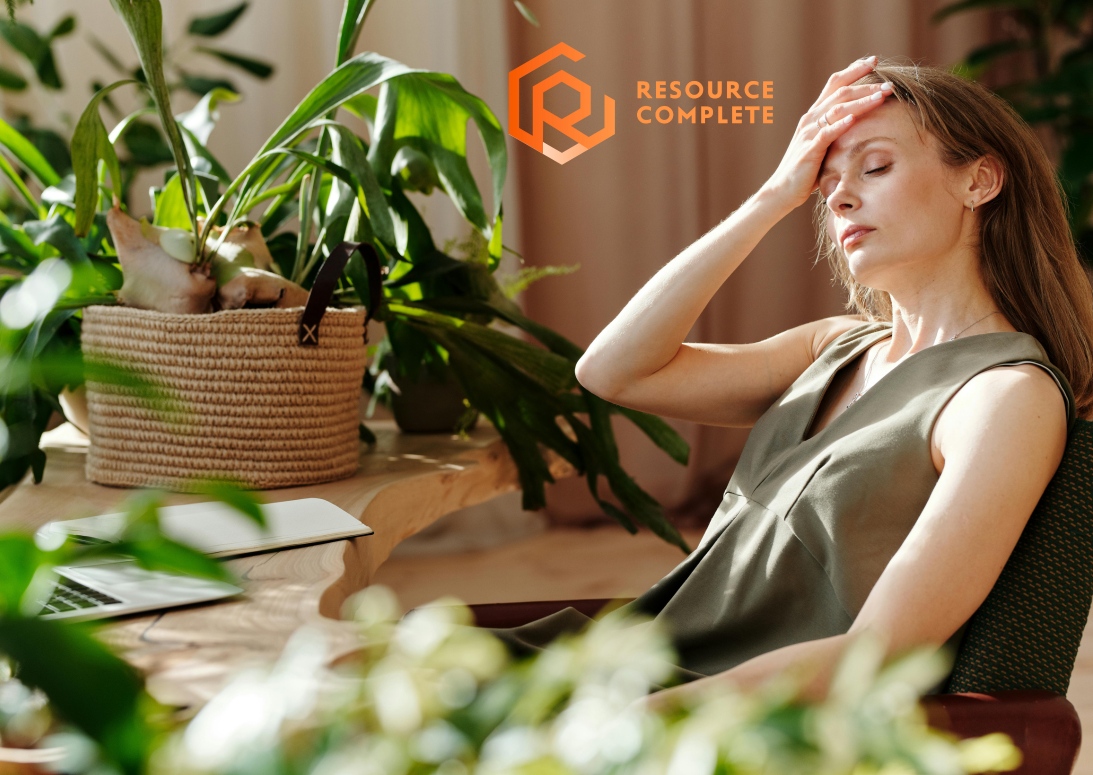 Stressed office worker resting head in hand beside laptop, Resource Complete logo in corner.
